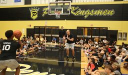 This is a photograph of an indoor basketball court where a group of young men are playing basketball. The court has a black